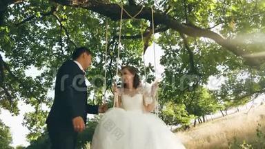 快乐的年轻夫妇新娘和新郎穿着婚纱在公园里荡秋千。 夏天在橡树枝上荡秋千